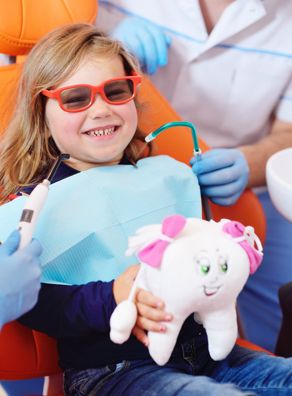 A baby girl in an orange dental chair smiles while holding a soft toy in the shape of a tooth. The dentist holds in his hands an ultraviolet curing light for polymerization of the seal.